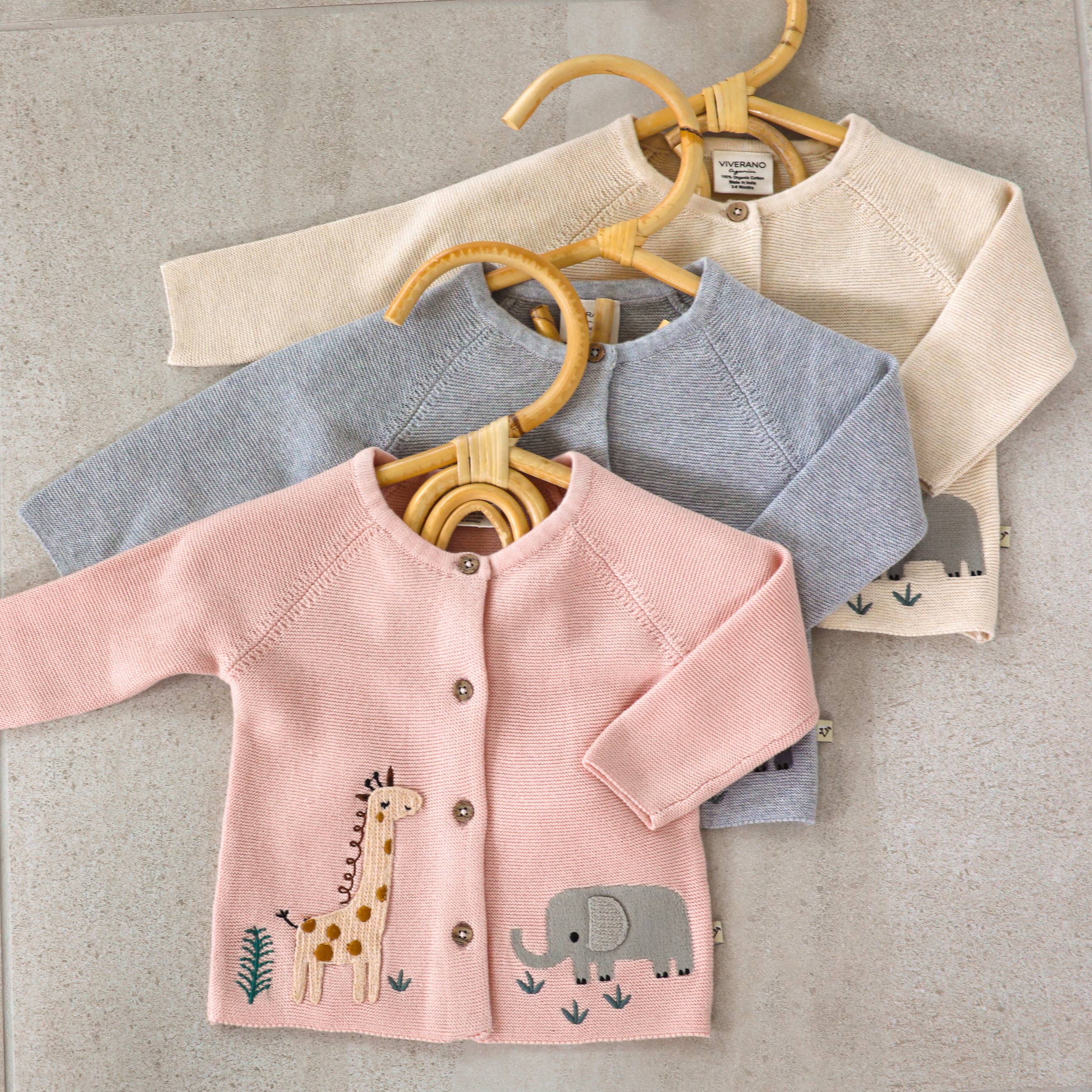 Three baby cardigans in pink, gray, and beige on hangers against a neutral background.