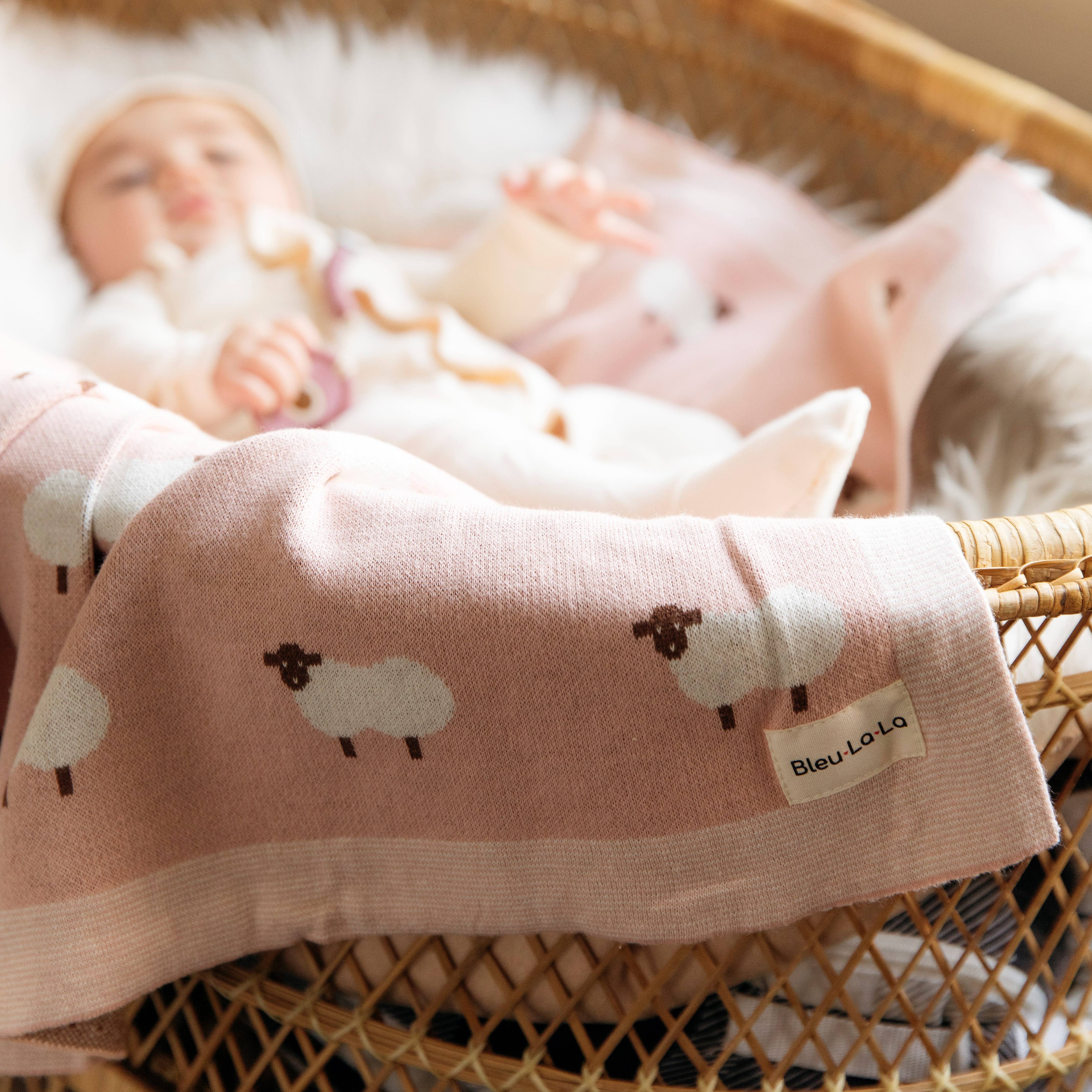Baby lying in a crib with a pink blanket featuring sheep patterns and a visible brand label.