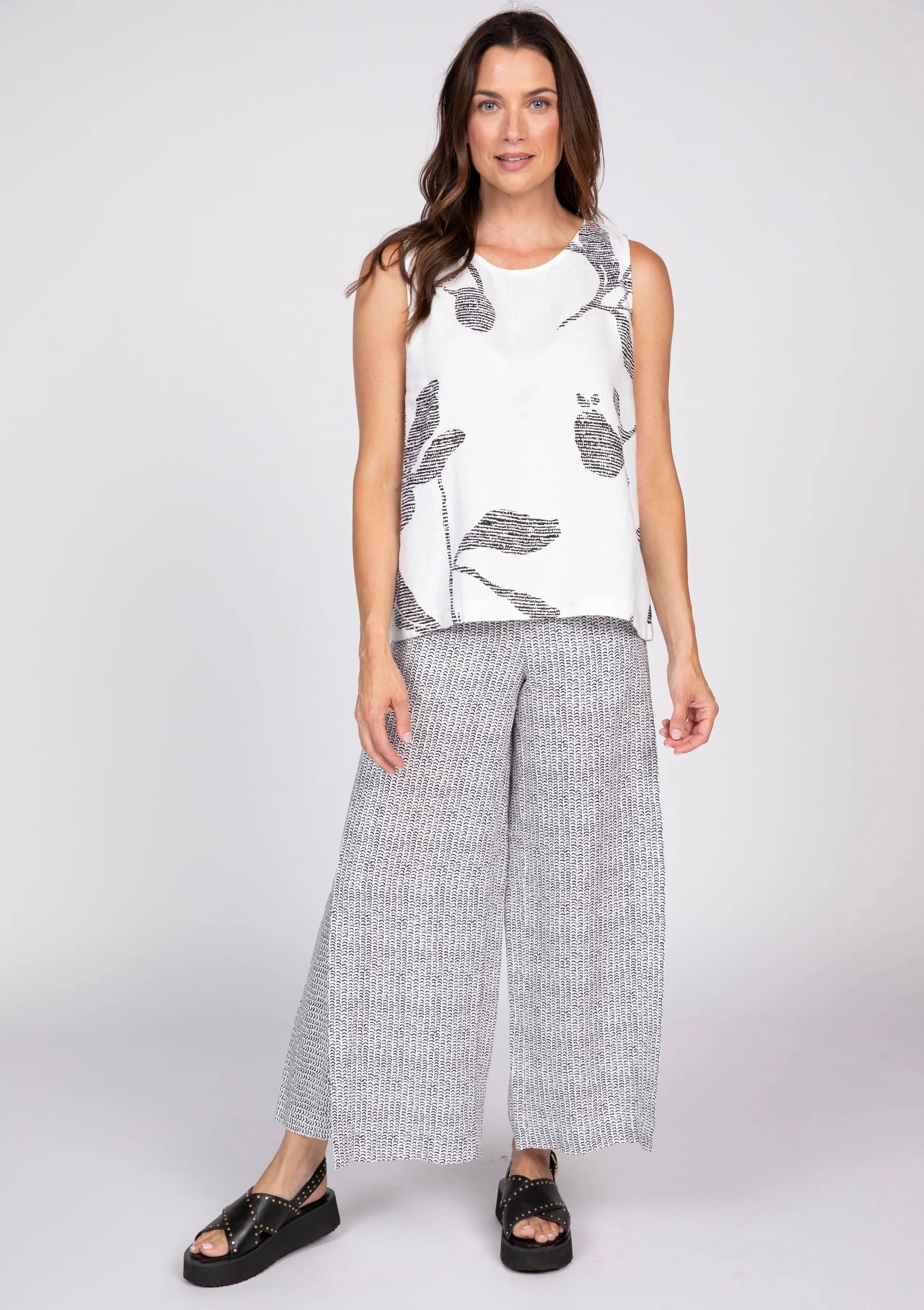 IMG_0133 — Women's white sleeveless tank top with black oversized floral bouquet print, front view, scoop neckline
IMG_0136 — Olivia women's white floral bouquet tank top, front torso view showing bold black botanical print
IMG_0135 — Women's white floral print tank top styled with black and white wide-leg pants and black platform sandals, full body front view
IMG_0134 — Women's white sleeveless tank top with large black leaf and floral print, back view
IMG_0137 — Close-up back view of women's white tank to
