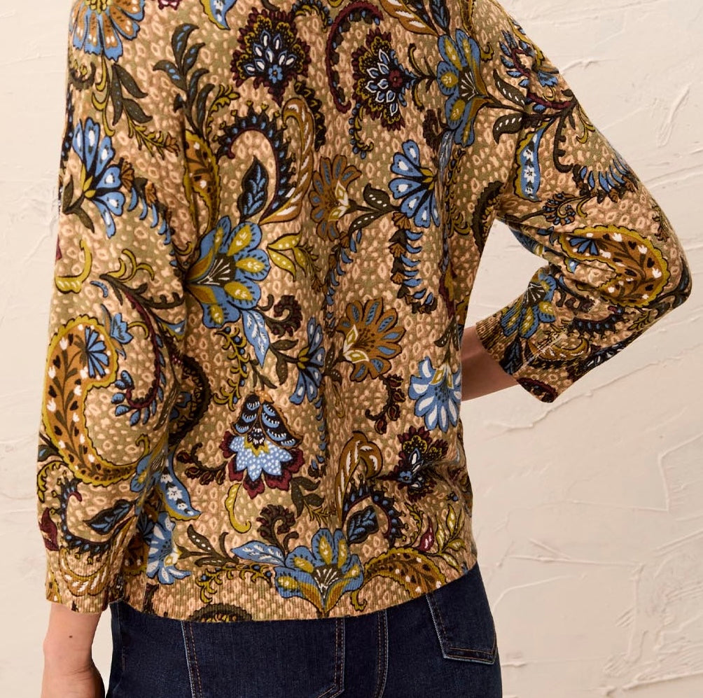 A close-up of a person wearing a long-sleeve blouse with a dense paisley and floral print in beige, blue, brown, and gold tones, paired with dark blue jeans against a light textured wall.