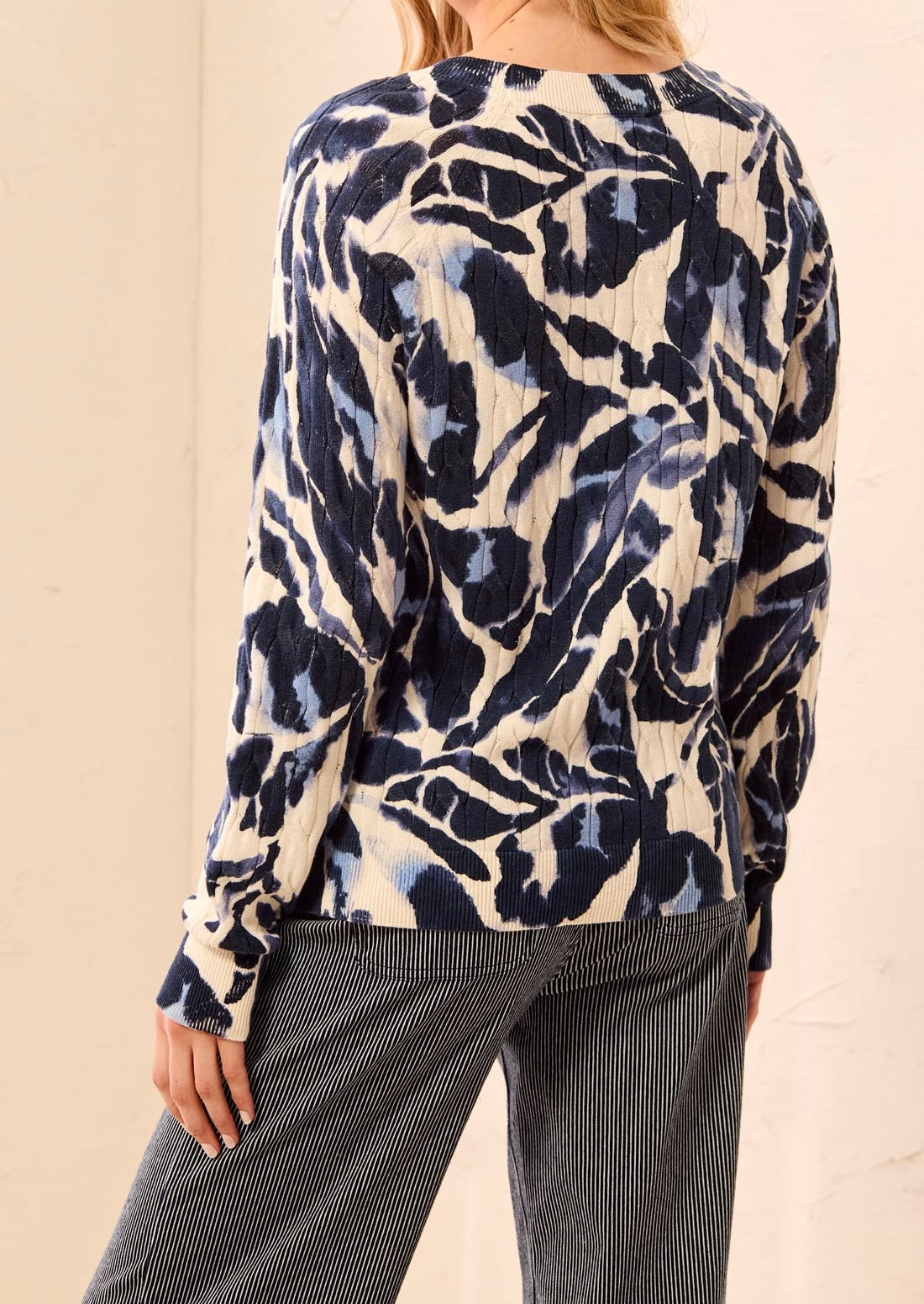 A woman shown from the back wearing a long-sleeve top with a navy and cream abstract pattern, paired with dark gray pinstriped pants, against a plain beige wall.