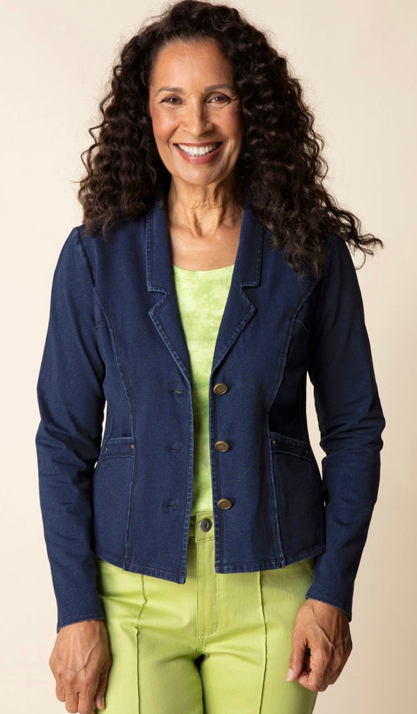 A smiling woman with long curly dark hair wears a navy blue button-front blazer over a light green top and matching green pants, standing against a plain beige studio background.
