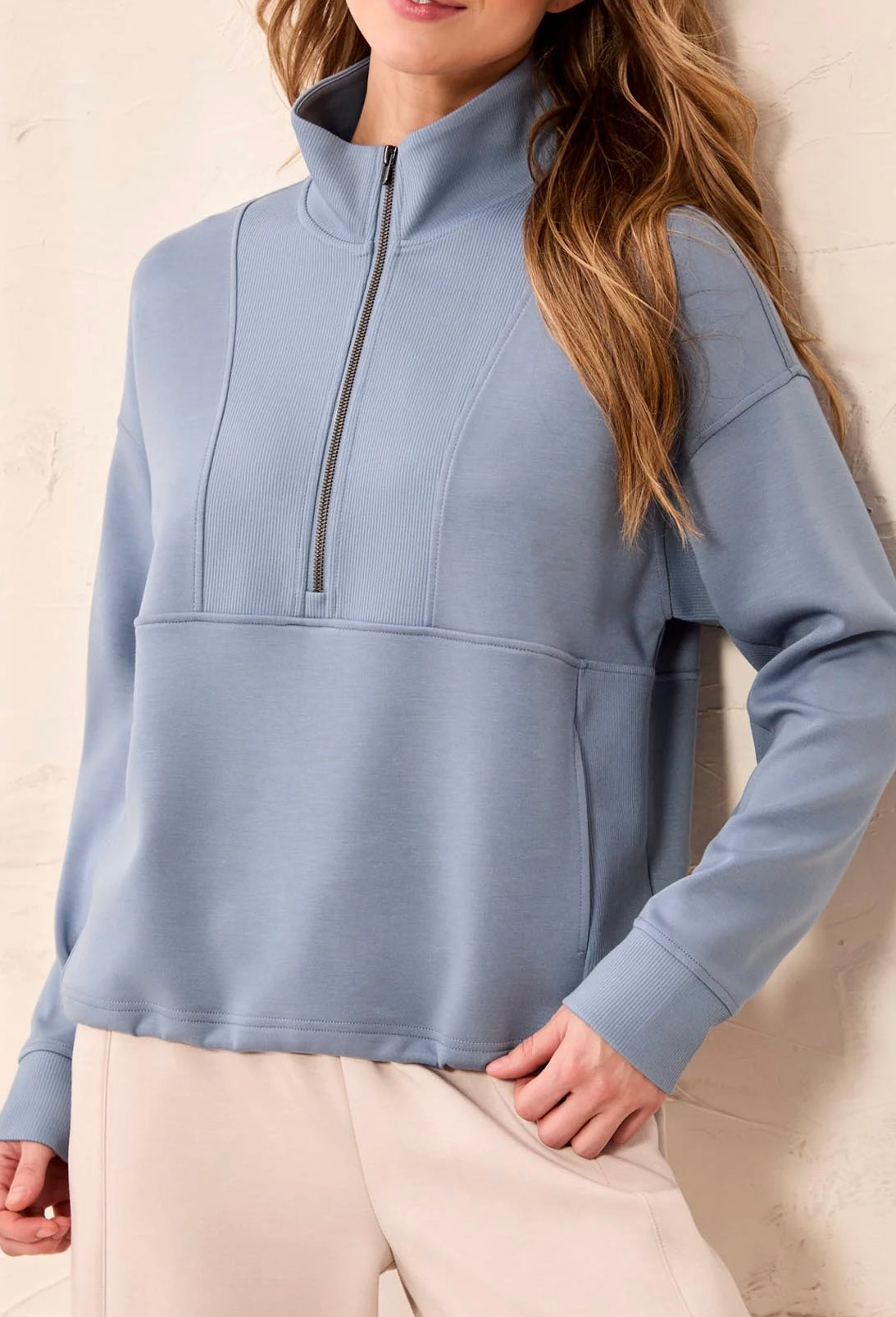 A woman wearing a dusty blue quarter-zip pullover sweatshirt with a mock neck and zippered chest pocket, paired with light beige pants, standing against a textured off-white wall.