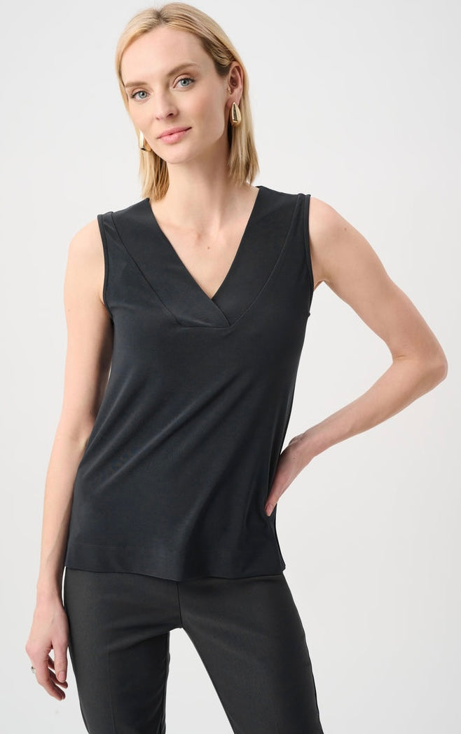 A woman modeling a sleeveless black V-neck top with a wrap-style front, paired with black pants, against a plain light gray studio background.