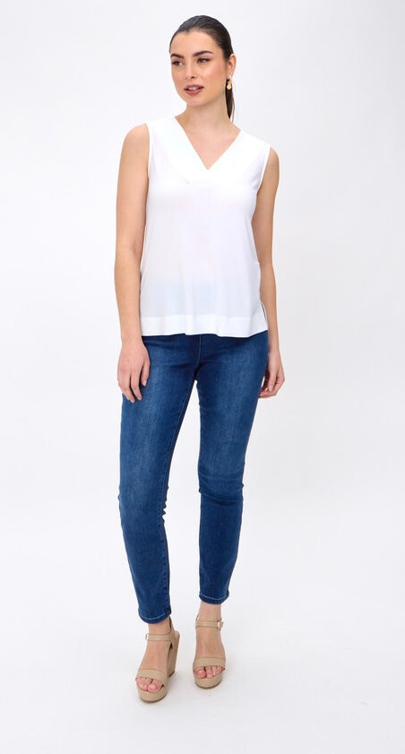 A woman modeling a sleeveless white V-neck top paired with dark blue skinny jeans and beige wedge sandals, standing against a plain white studio background.
