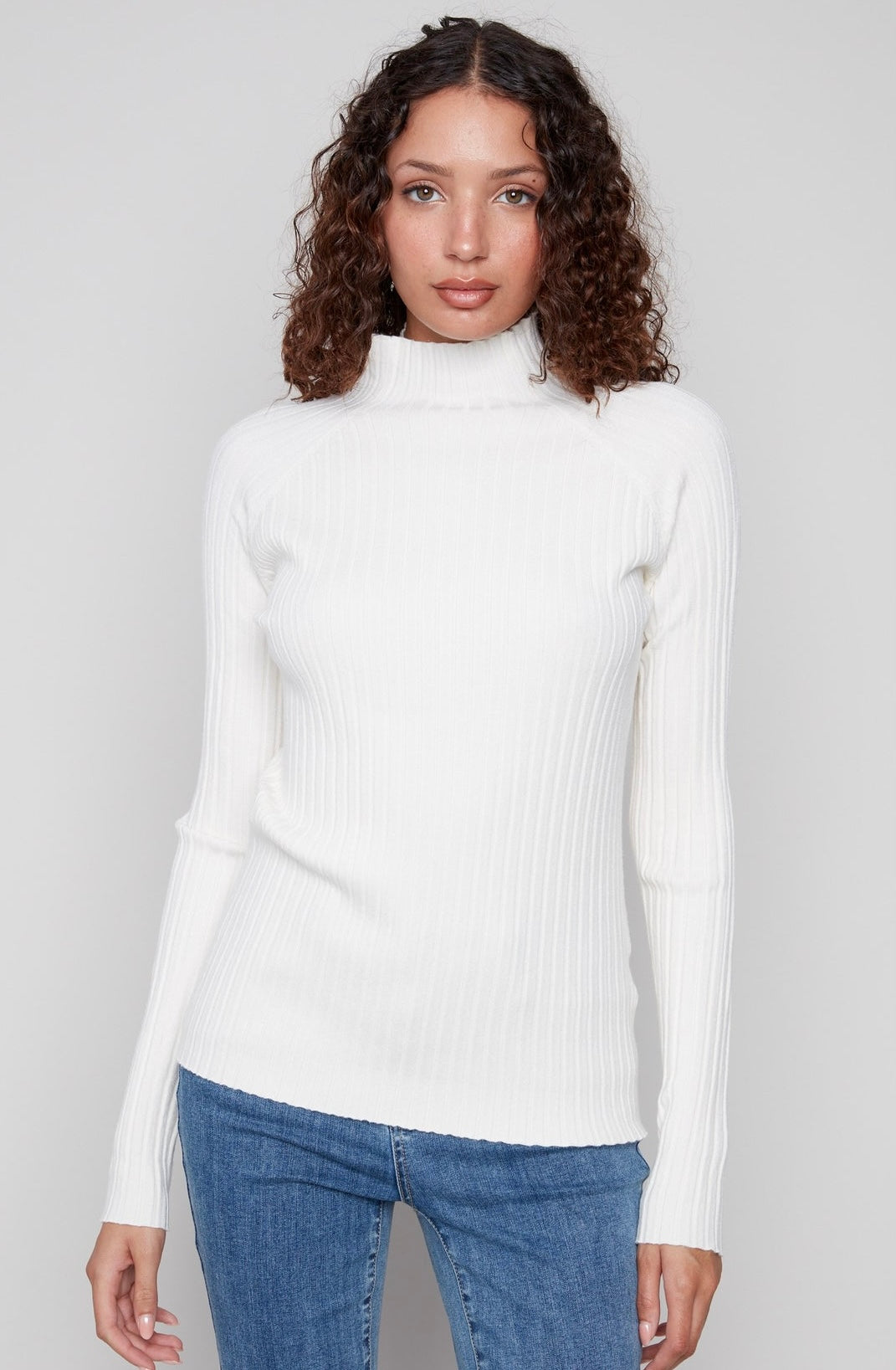 A woman with curly brown hair wearing a white ribbed mock neck long-sleeve sweater and blue jeans, standing against a light gray studio background.
