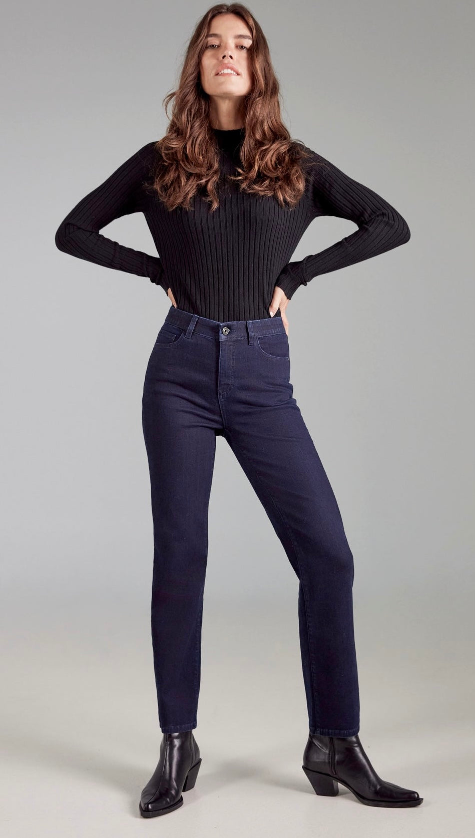 A woman modeling a black ribbed long-sleeve mock-neck top and dark navy blue skinny jeans with black ankle boots, standing against a light gray studio background.
