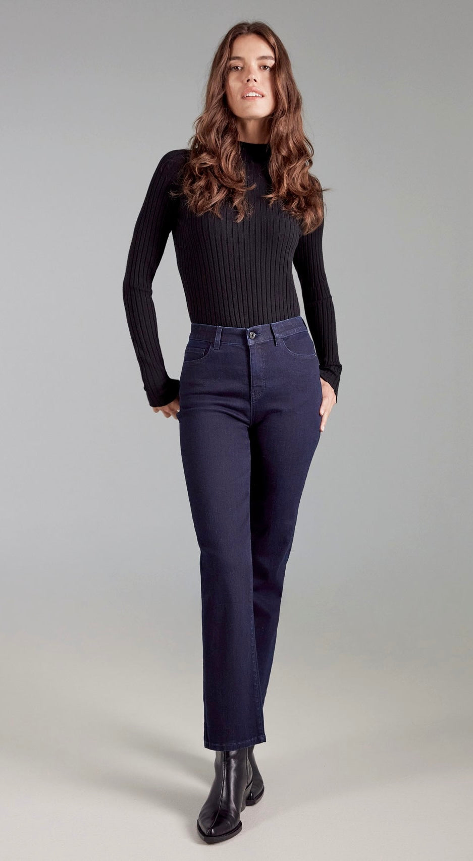 A woman modeling a black ribbed long-sleeve mock neck top and navy blue straight-leg jeans, paired with black leather ankle boots, against a solid light gray studio background.