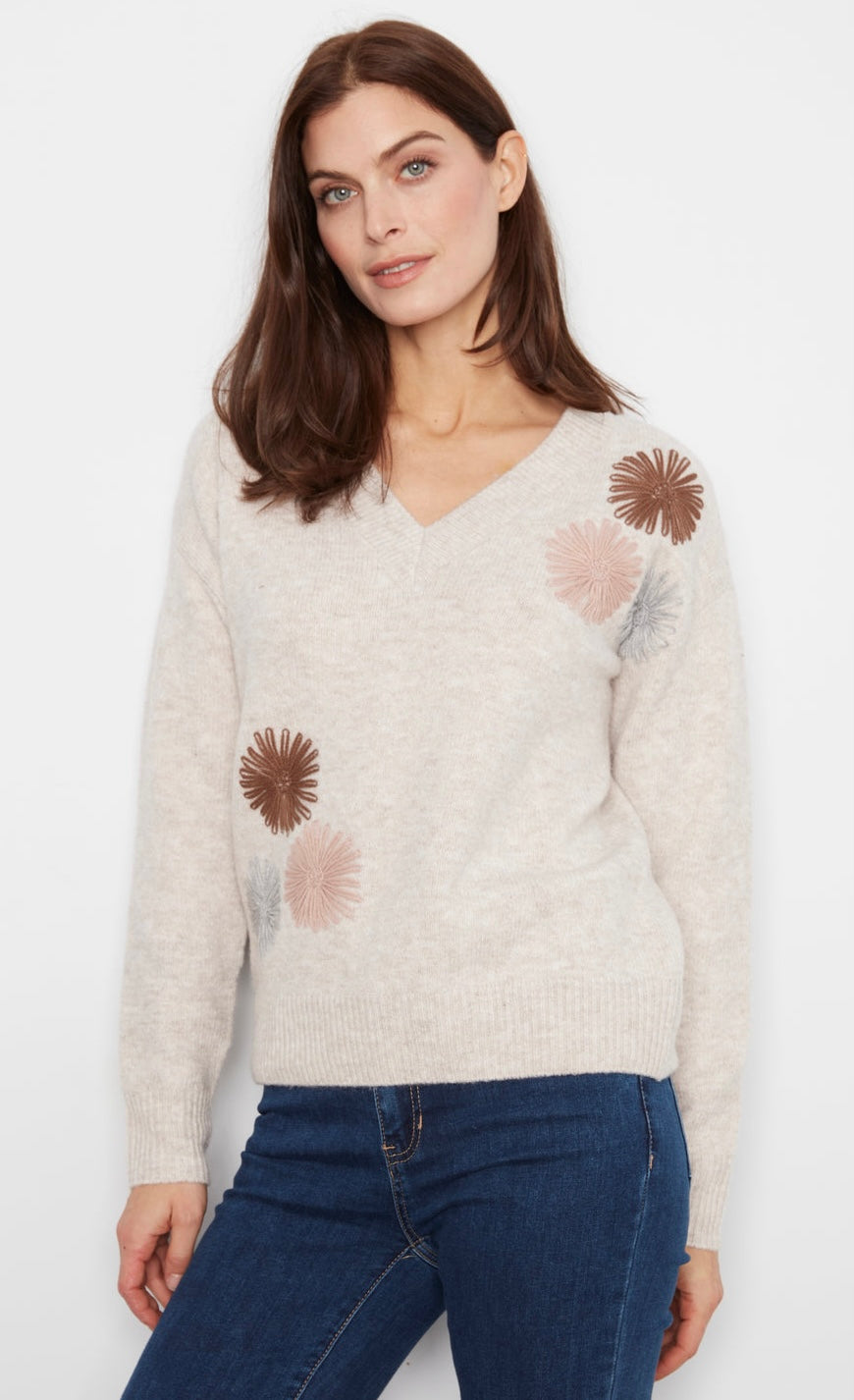 A woman stands against a white wall wearing a cream knit sweater with brown and pink floral patches, dark blue skinny jeans, and black ankle boots.