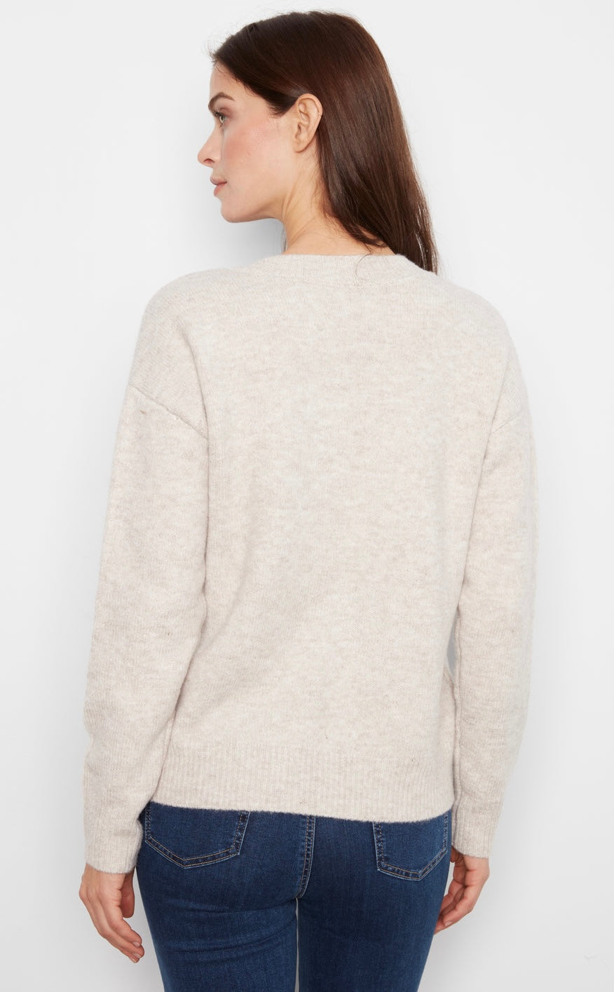 A woman shown from the back wearing a light beige knit sweater and dark blue jeans against a plain white background.