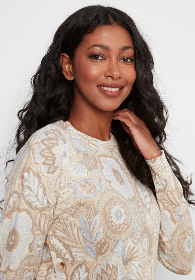 A woman with long, wavy black hair is shown from the chest up, wearing a beige and white patterned knit sweater against a plain white studio background.