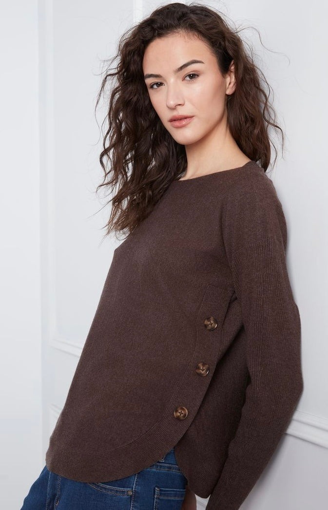 A woman wearing a brown knit sweater with a boat neckline and three decorative buttons on the side, paired with blue jeans, set against a white paneled wall.