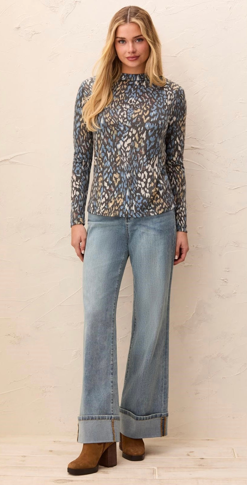 A woman modeling a long-sleeve mock neck top with a blue, gray, and beige animal print, paired with light-wash wide-leg jeans and brown boots, standing against a textured beige wall.