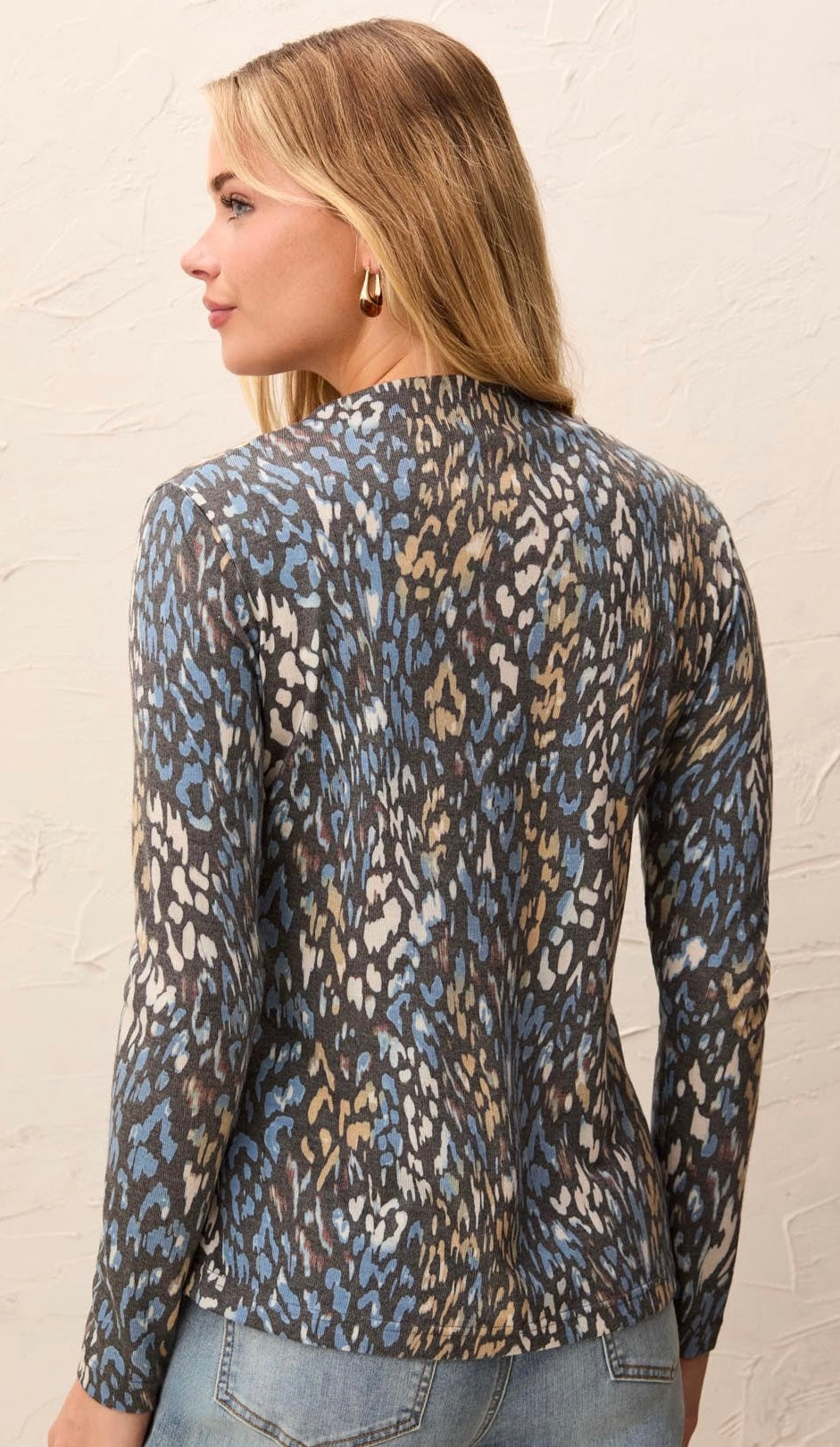 A woman shown from the back wearing a long-sleeve leopard print top with blue, beige, and gray tones, paired with light-wash jeans against a textured off-white wall.