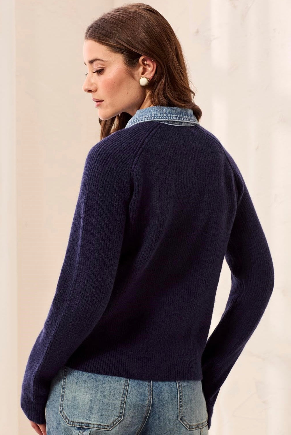A woman shown from the back wearing a dark navy ribbed knit cardigan layered over a light blue denim shirt, paired with blue jeans against a neutral cream-colored background.