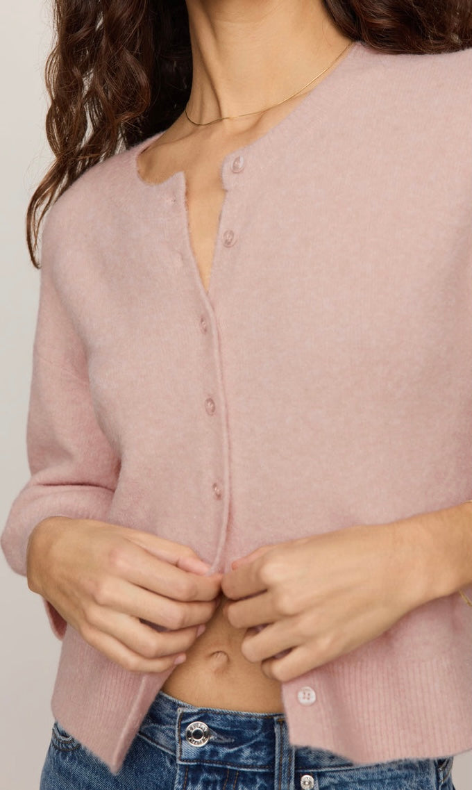 A close-up of a woman wearing a dusty pink knit cardigan with a buttoned front, paired with blue jeans.