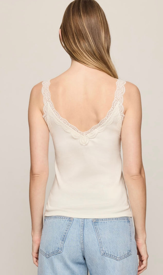 A woman viewed from the back wearing a cream-colored lace-trimmed camisole top and light blue jeans, set against a plain light beige studio background.