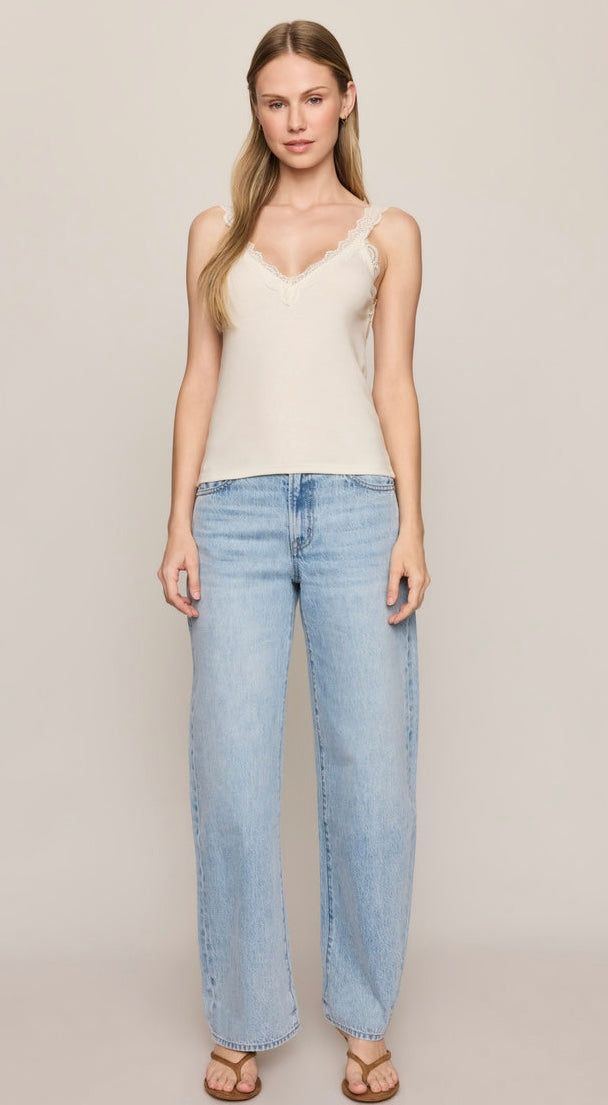 A woman modeling a cream-colored lace-trimmed camisole top paired with light-wash wide-leg jeans and brown sandals, set against a plain beige studio background.