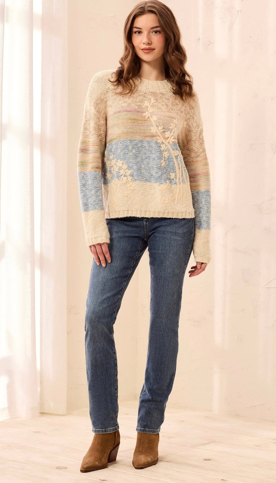 A woman modeling a cream and light blue knit sweater with floral embroidery, paired with blue jeans and brown ankle boots, standing in a brightly lit room with white curtains.