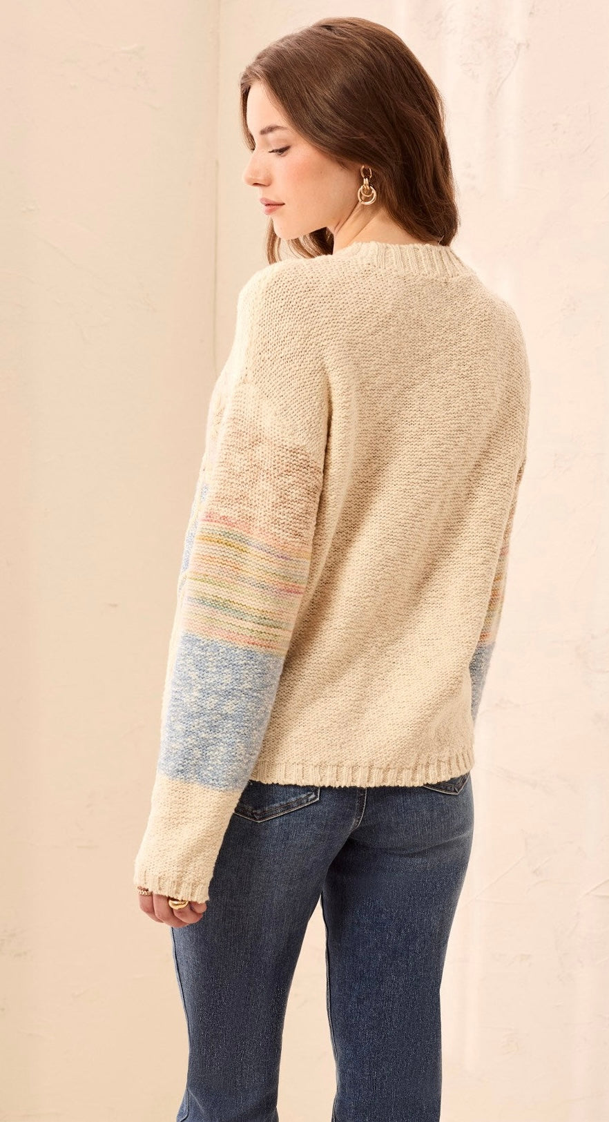 A woman wearing a cream-colored knit sweater with pastel striped accents on the sleeves, paired with dark blue jeans, shown from a side-back angle against a neutral beige background.