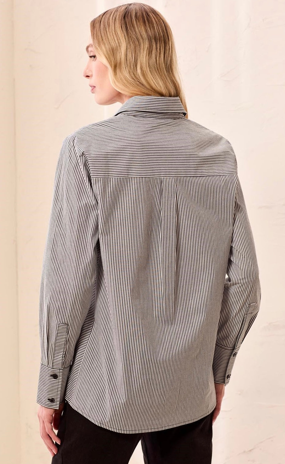 A woman shown from the back wearing a gray and white vertically striped button-down shirt with a spread collar and dark pants, set against a light beige background.