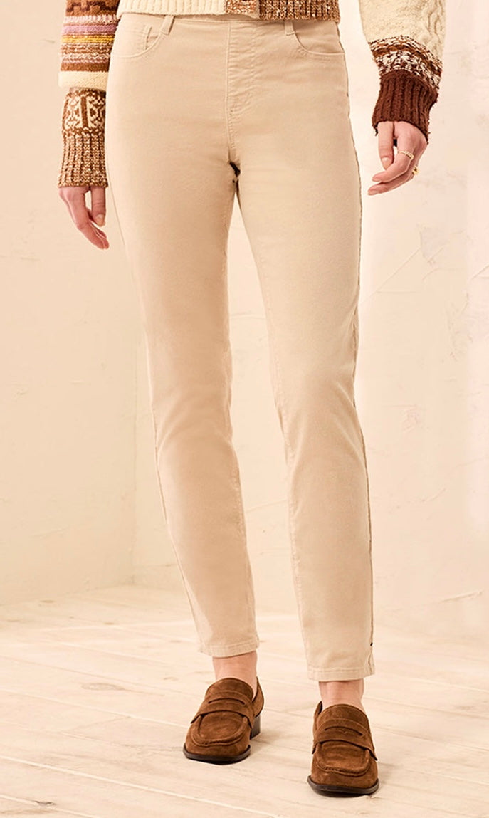 A person wearing slim-fit beige pants, a cream and brown knit sweater, and brown suede loafers, standing against a light beige wall and wood floor.