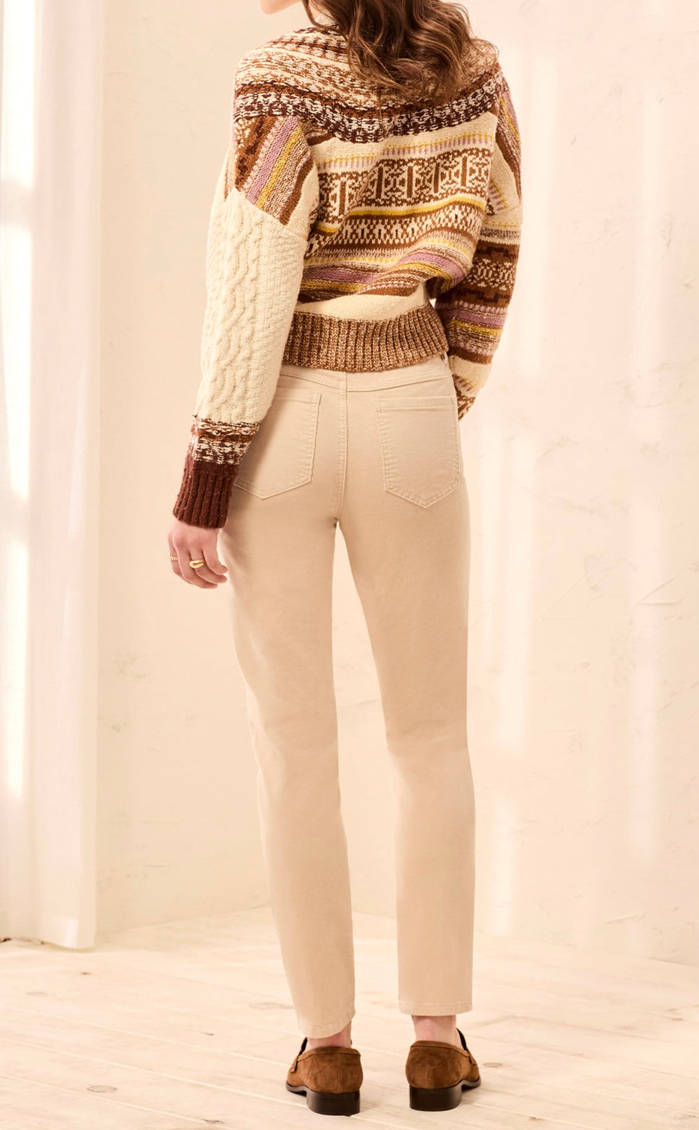 A woman shown from the back wearing a cream, brown, and tan Fair Isle knit sweater with cable-knit sleeves, paired with beige straight-leg pants and brown suede loafers, in a brightly lit indoor setting.