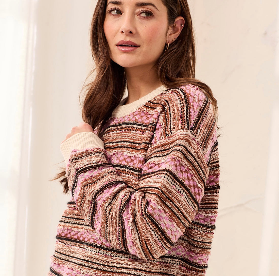 A woman wearing a chunky knit sweater with horizontal stripes in pink, brown, beige, and black, posed against a soft, light-colored indoor background.