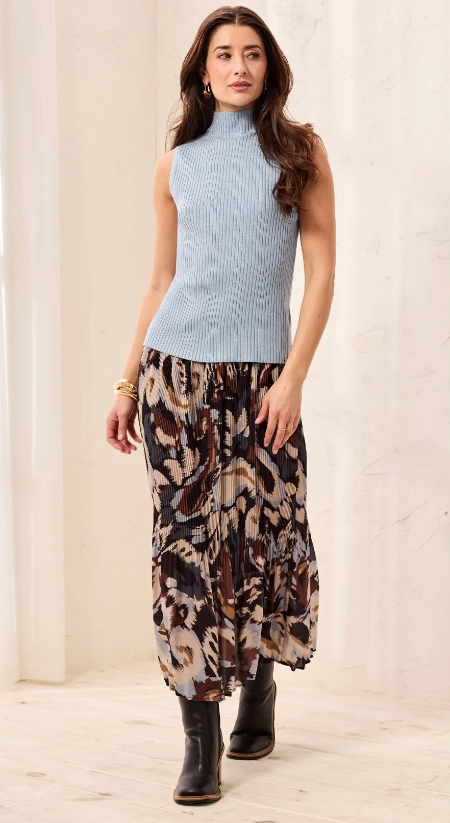 A woman modeling a sleeveless light blue ribbed mock-neck top paired with a brown, beige, and black abstract print maxi skirt and black ankle boots, standing against a light beige textured wall.