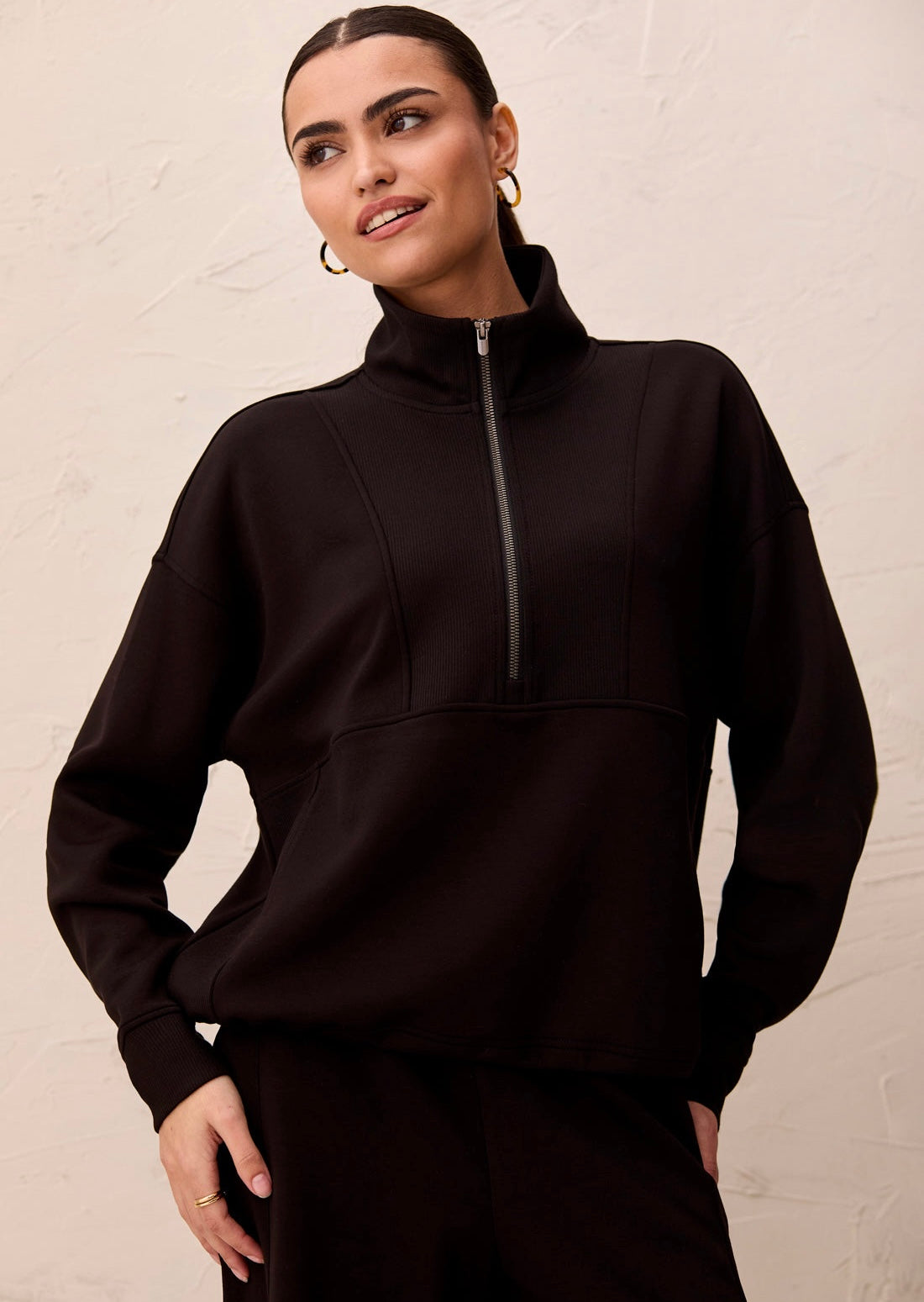 A woman wearing a black mock-neck quarter-zip sweatshirt and matching black pants, standing against a textured off-white wall.