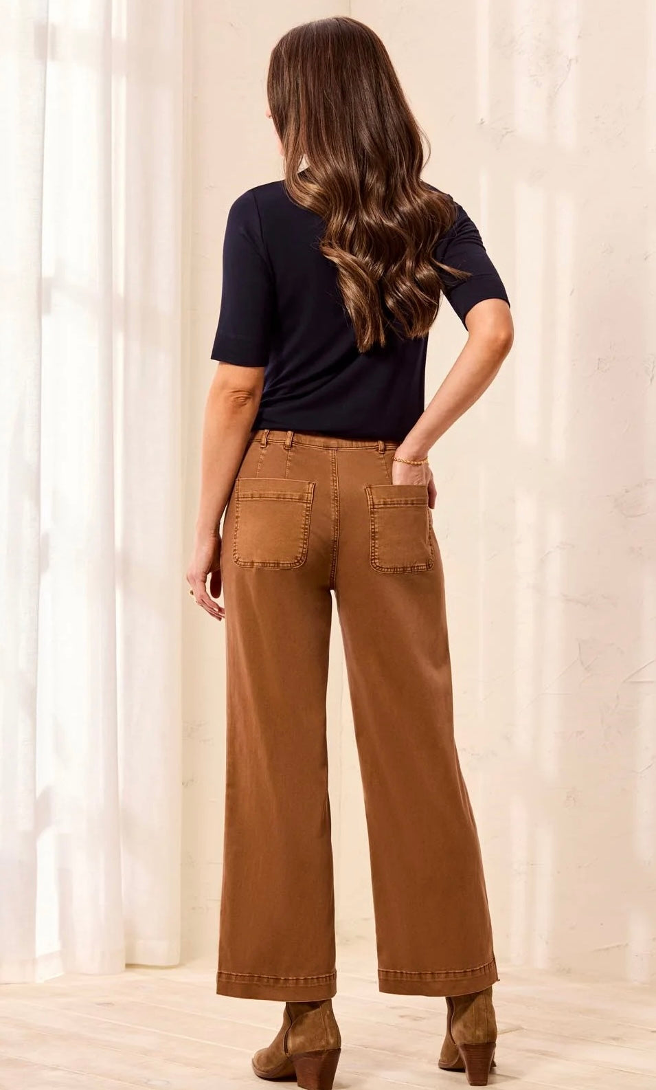 A woman shown from the back wearing brown wide-leg cropped pants, a dark navy short-sleeve top, and brown heeled boots in a bright indoor setting with white curtains.