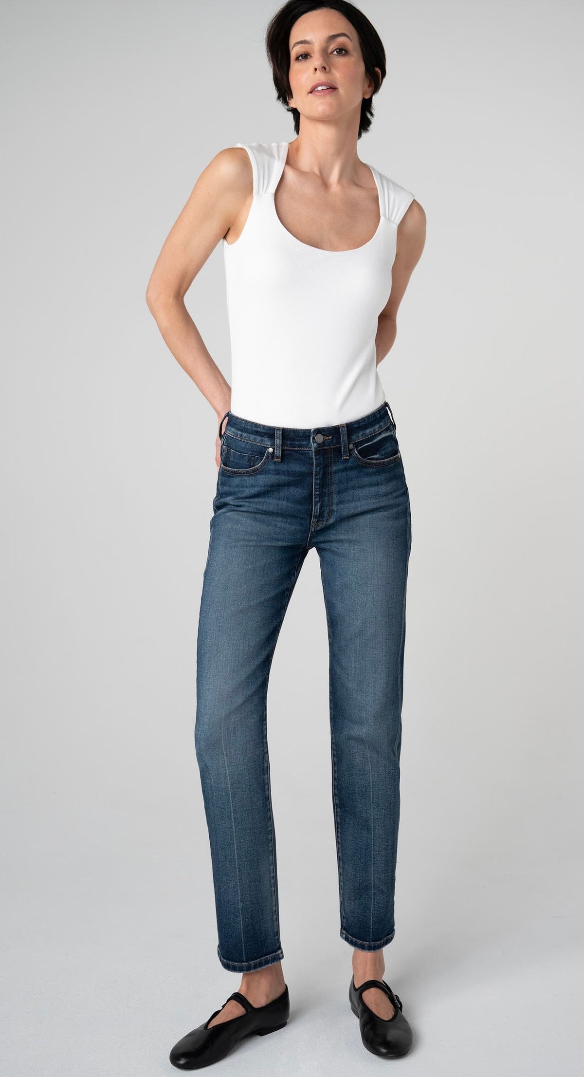 A woman modeling a pair of dark wash blue jeans with a white tank top and black flat shoes, standing against a light gray studio background.
