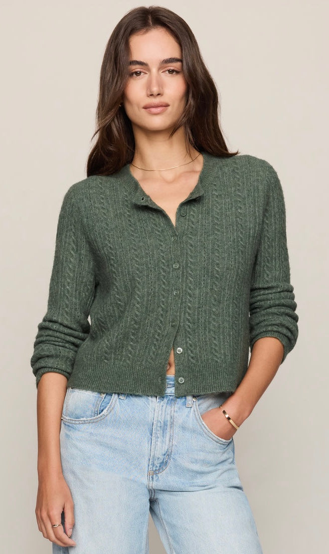 A woman wearing a sage green cable-knit button-up cardigan sweater and light-wash blue jeans, standing against a solid light beige background.