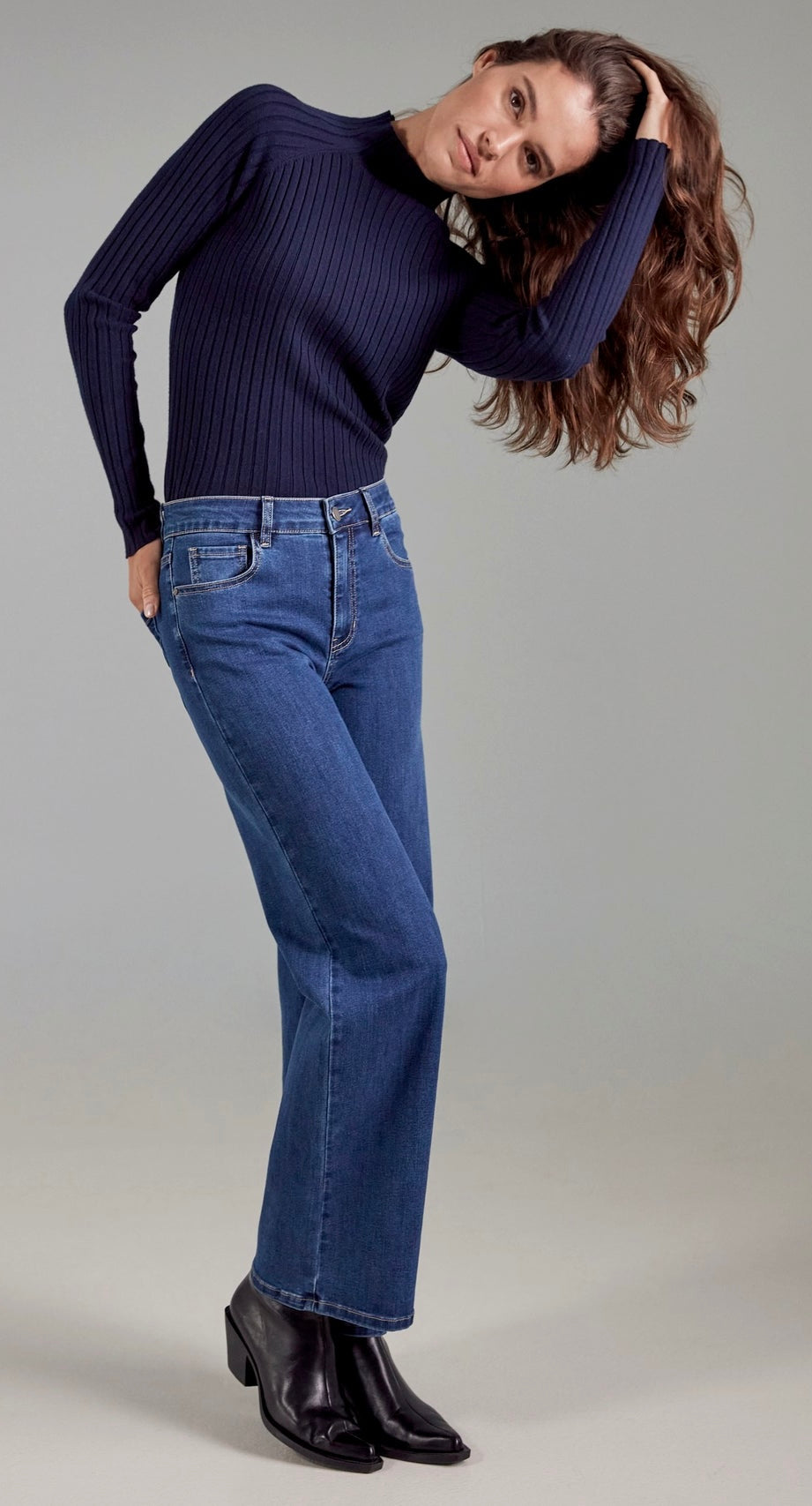 A woman modeling a dark navy ribbed long-sleeve mock neck sweater and blue jeans with black ankle boots, posed against a solid light gray studio background.