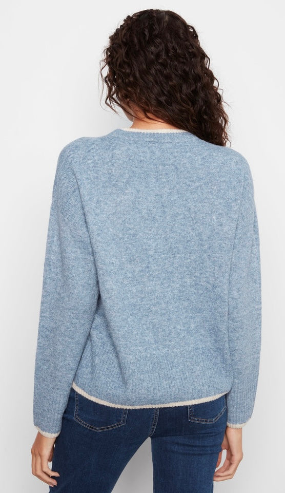 A woman viewed from the back wearing a light heathered blue knit sweater with cream ribbed trim at the collar and hem, paired with dark blue jeans against a plain white background.