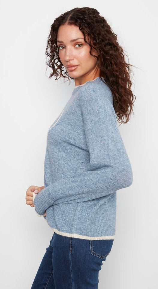 A woman with curly brown hair wearing a light blue heathered knit sweater and dark blue jeans, shown from a side-back angle against a plain white studio background.