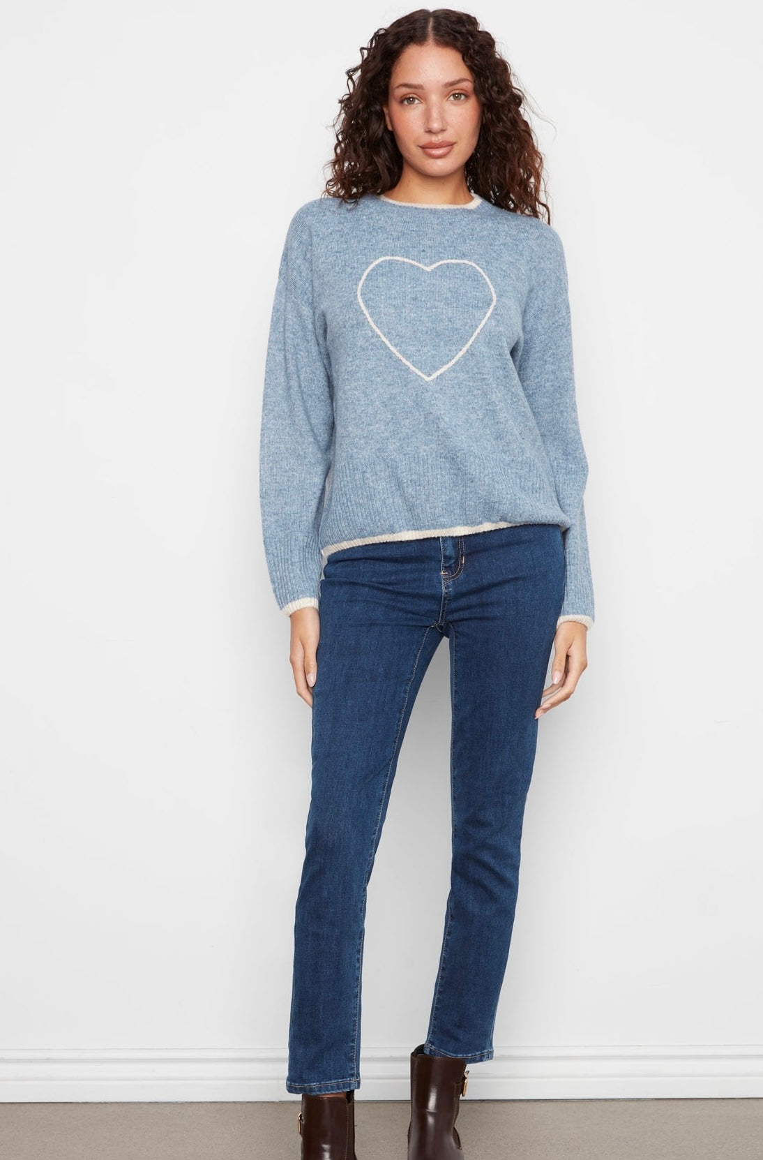 A woman modeling a light blue heathered knit sweater with a white heart outline on the chest, paired with dark blue jeans and brown ankle boots, standing against a white wall.