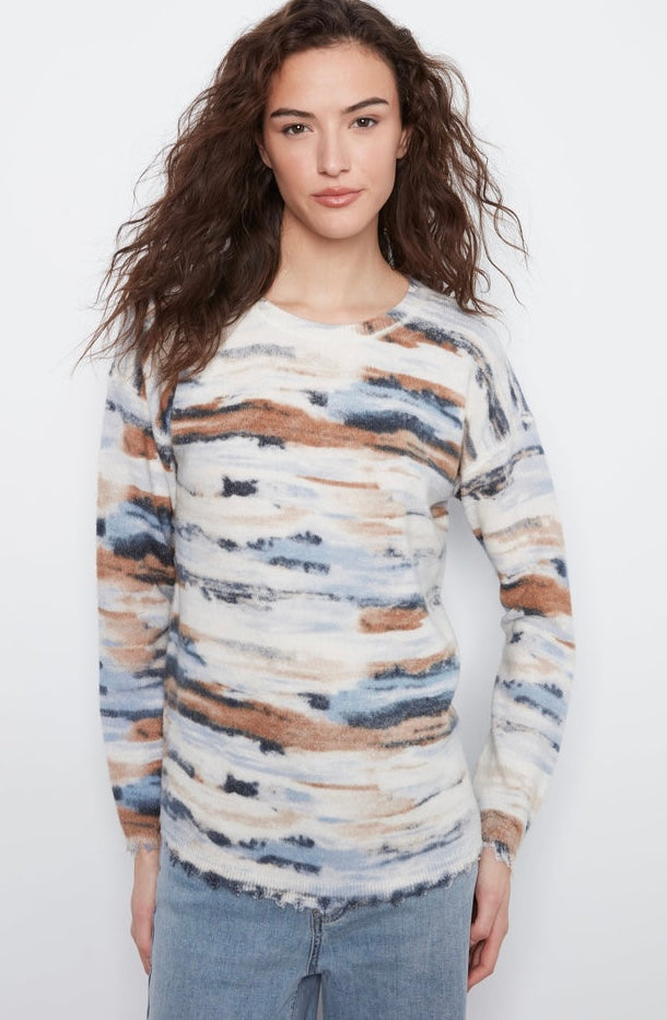 A woman modeling a long-sleeve knit sweater with an abstract watercolor pattern in beige, brown, blue, and white, paired with light-wash jeans against a plain white background.