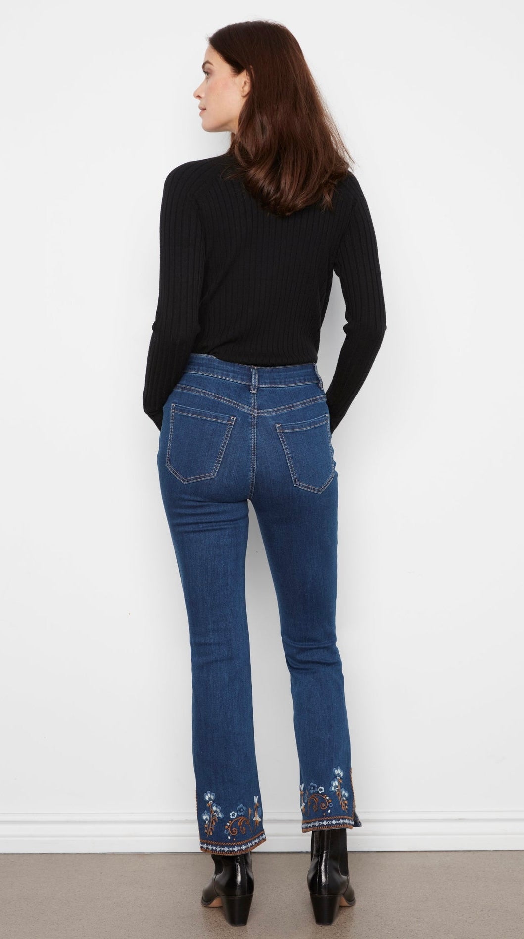 Person wearing blue jeans with floral embroidery at the bottom, standing against a white wall.