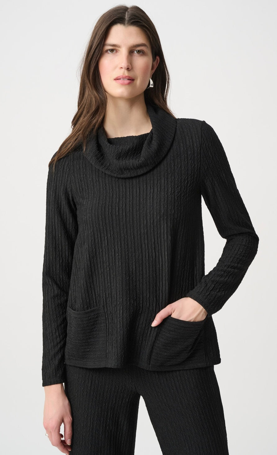 A woman wearing a black ribbed knit cowl-neck sweater with long sleeves and front patch pockets, paired with matching black ribbed pants, against a light gray studio background.