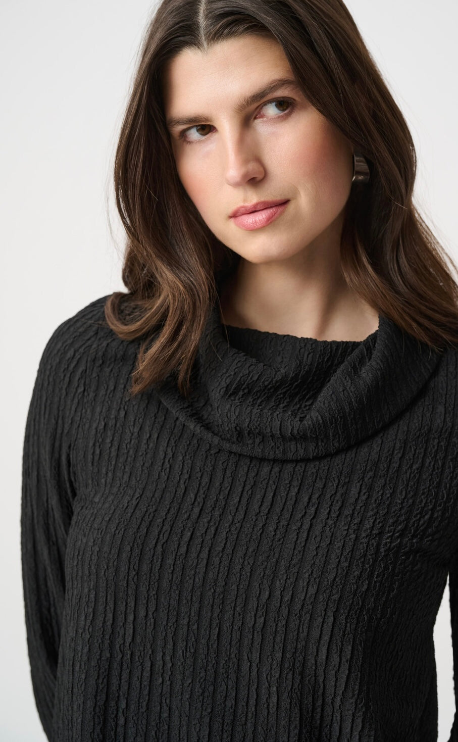 A woman wearing a black ribbed cowl-neck sweater, shown from the chest up against a plain light gray studio background.