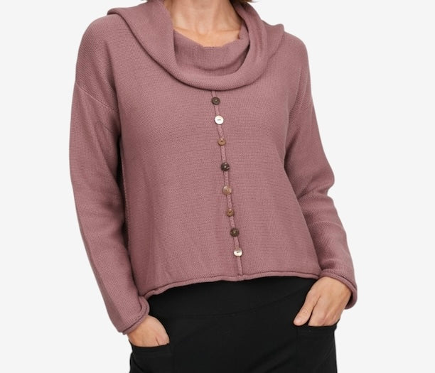 A woman wearing a dusty rose cowl-neck knit sweater with a vertical row of decorative buttons down the front, paired with black pants, against a light gray background.