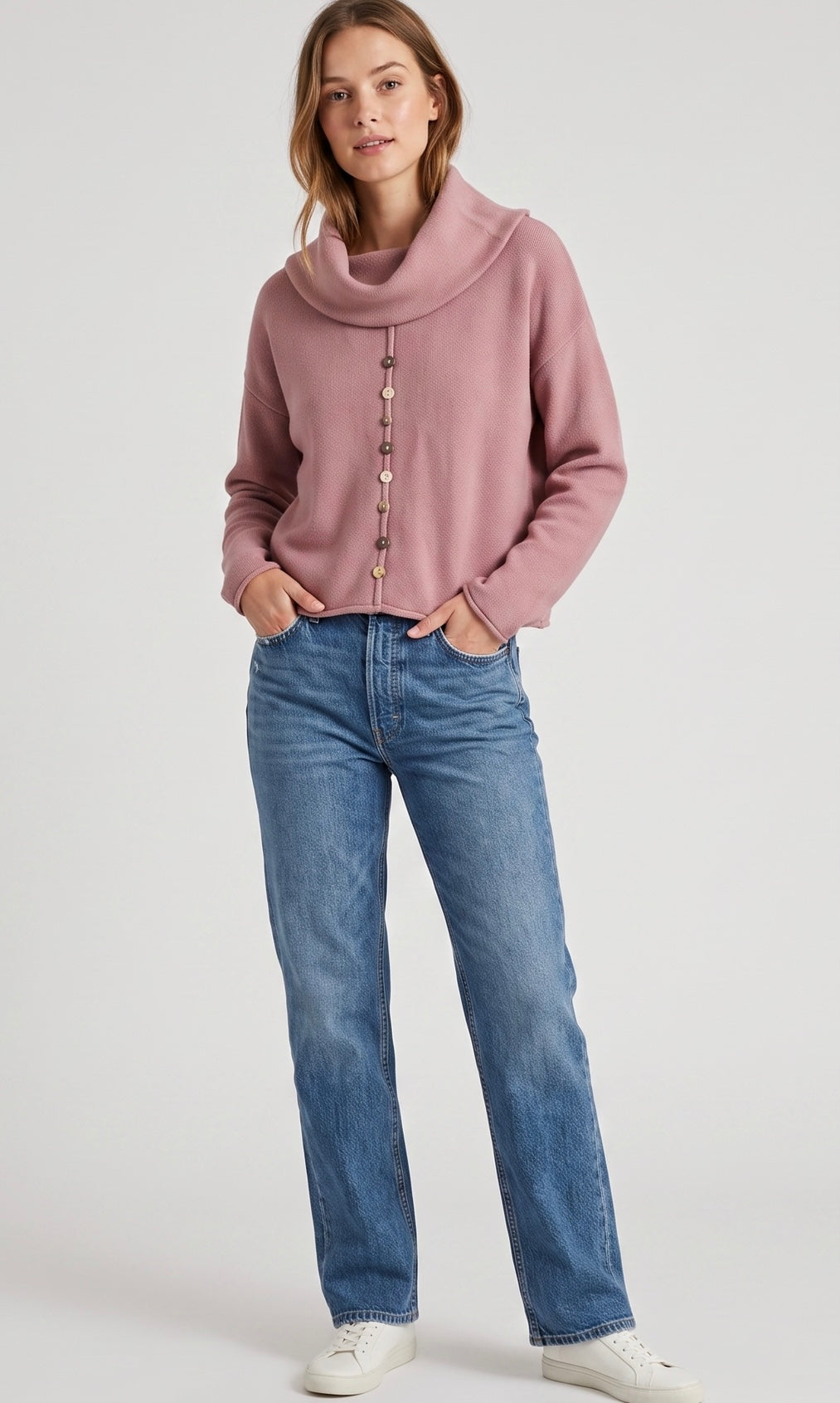 A woman modeling a dusty rose cowl-neck knit sweater with decorative buttons down the front, paired with blue jeans and white sneakers, against a light gray studio background.