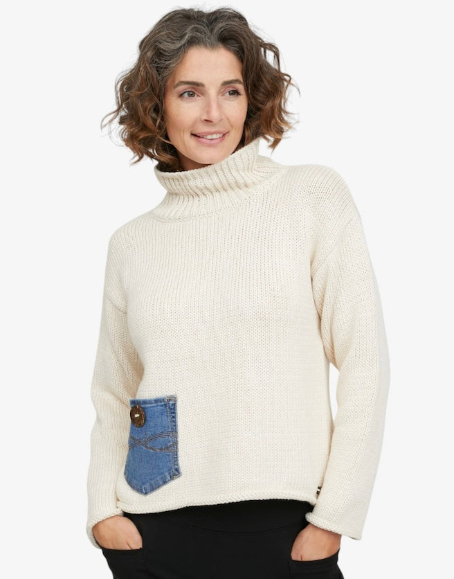 A woman wearing a cream-colored knit turtleneck sweater with a blue denim pocket detail on the front, paired with black pants, against a plain white background.