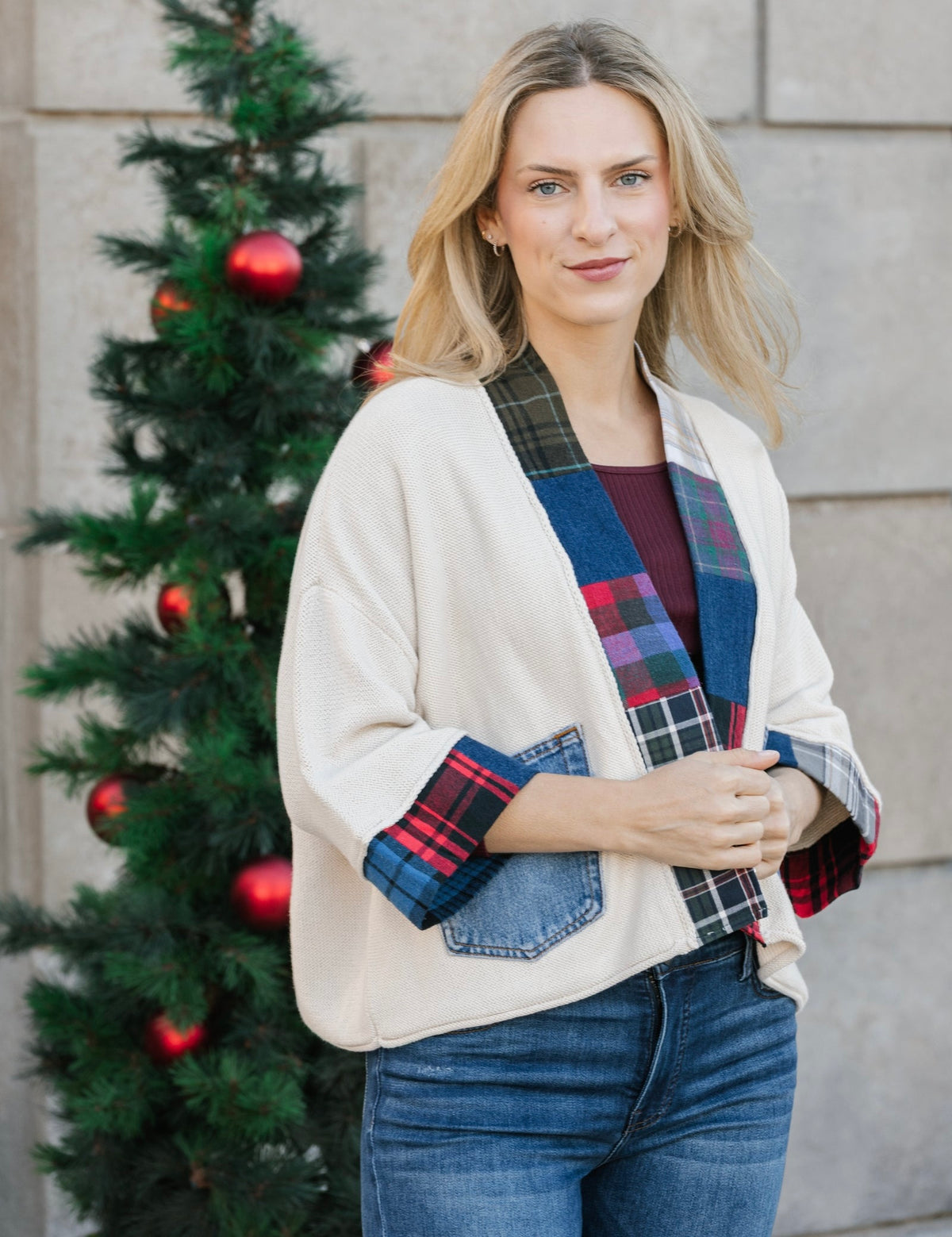 A woman wearing a cream-colored cardigan with blue denim sleeves and colorful plaid patchwork details, standing in front of a stone wall and a decorated Christmas tree.
