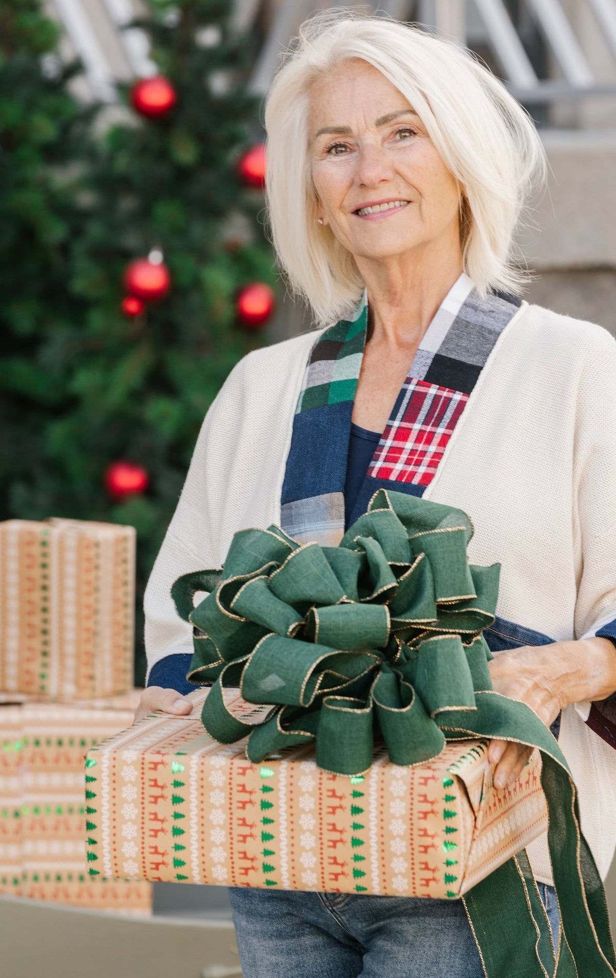 A smiling older woman with short white hair holds a large wrapped gift box with a decorative green bow. She wears a cream cardigan over a plaid shirt and jeans, standing in front of a blurred Christmas tree with red ornaments.