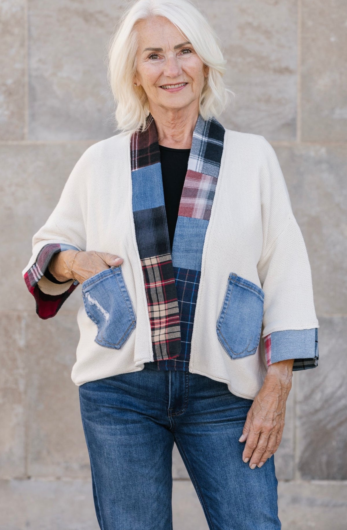 A smiling older woman stands outdoors against a stone wall wearing a cream open-front cardigan with blue denim patch pockets and a multicolor plaid collar, paired with blue jeans.