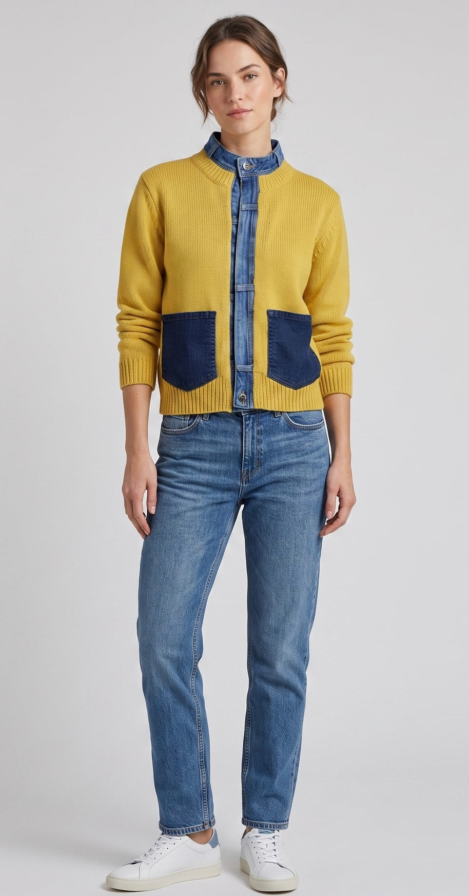 A model wearing a mustard yellow cardigan with dark blue denim pockets and a denim collar, paired with blue jeans and white sneakers, against a light gray studio background.