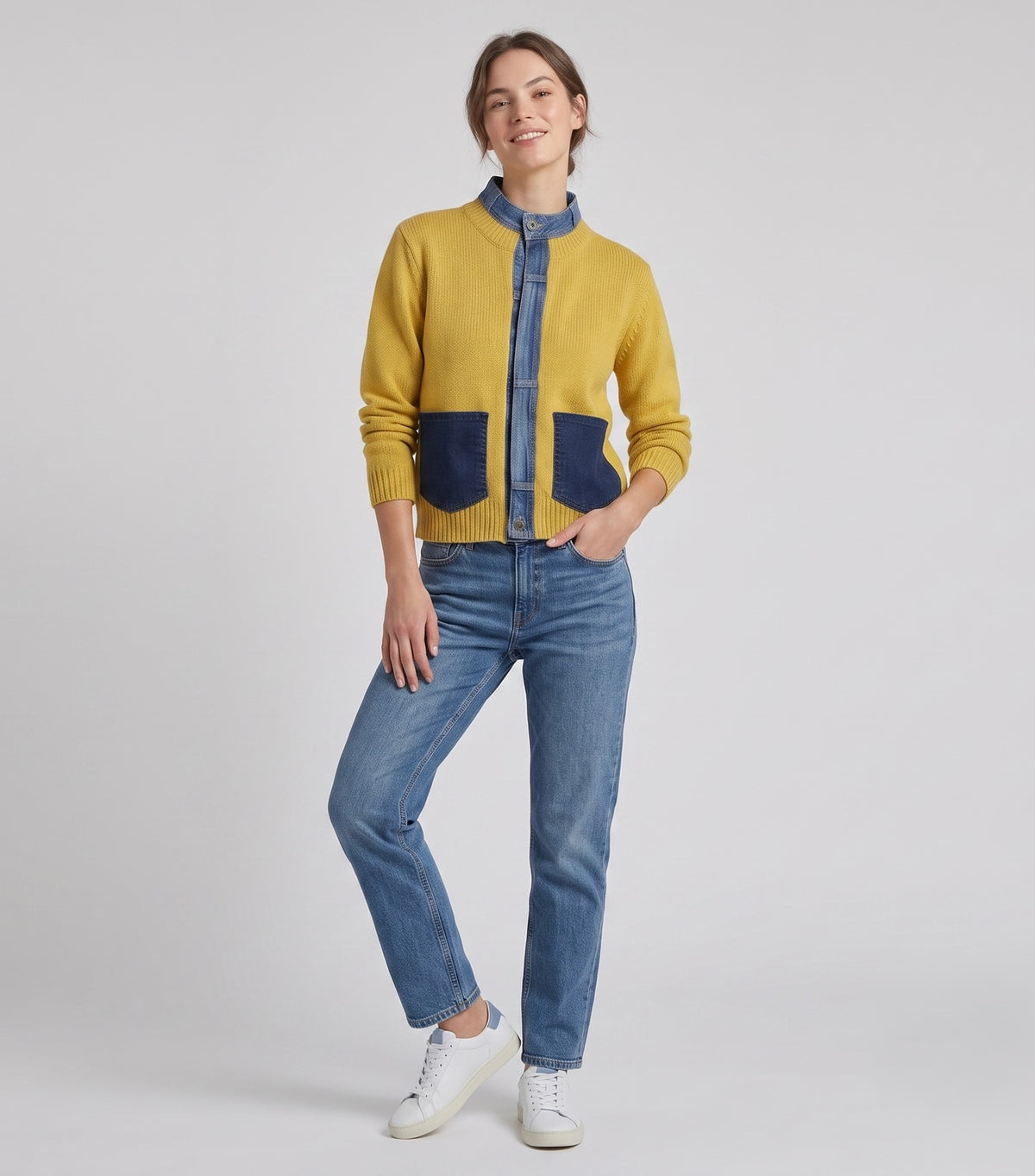 A woman modeling a mustard yellow and blue color-blocked cardigan with denim-style trim, paired with blue jeans and white sneakers, against a light gray studio background.