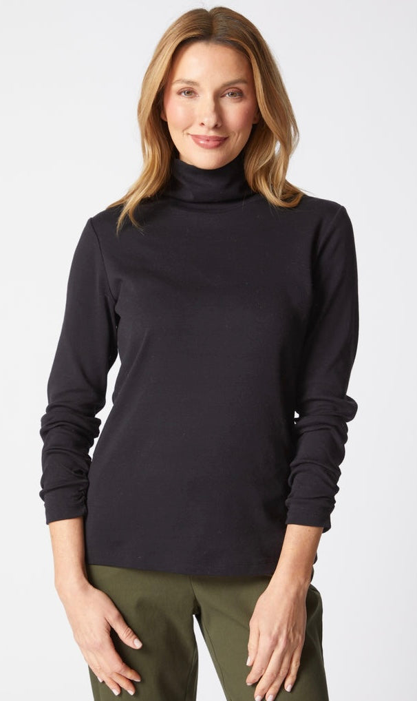 A woman wearing a black long-sleeve turtleneck sweater and olive green pants, standing against a light gray studio background.